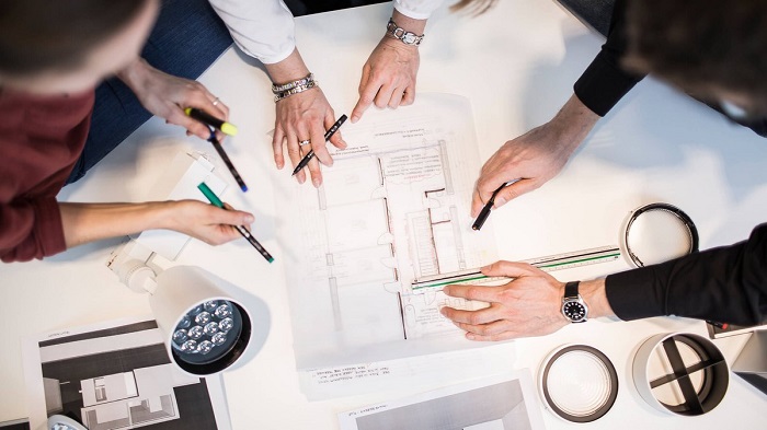 Three people are discussing a design laid out on the table in front of them. Their faces are not seen. Two of them have pens in their hands while the third one has a pen and ruler.